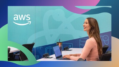 一名女子坐在教室的桌子前,面前放着笔记本电脑和一个水瓶。她微笑着看着远方。她身后是一面蓝色的大墙,上面写着“AWS”。