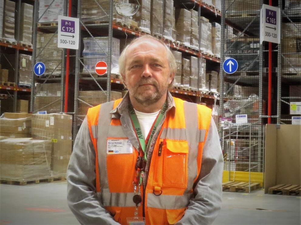 Oberkörperprofil eines Amazon Mitarbeiters in orange-farbener Sicherheitsweste. Er steht vor einem Hochregallager.