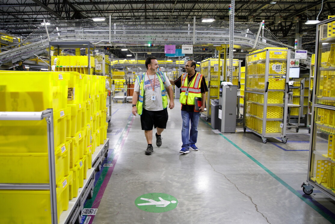 Two men walk along an aisle in a large warehouse space. 