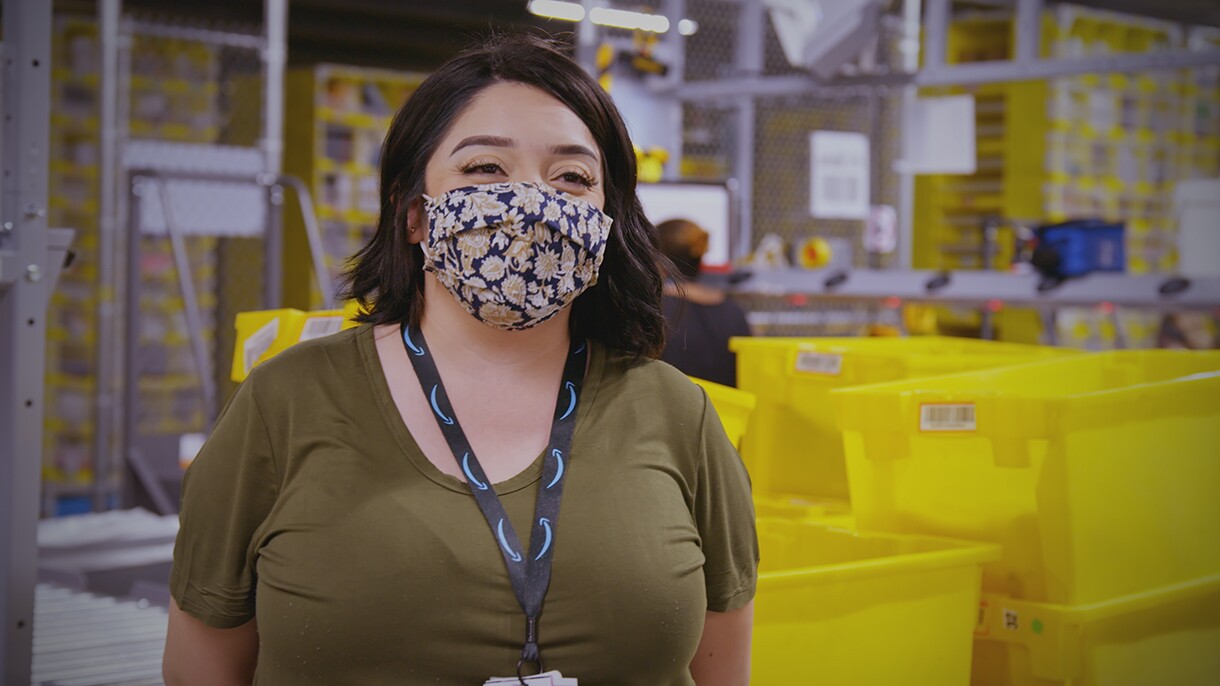 A woman wearing a t-shirt, lanyard, and face mask stands in an Amazon fulfillment center.