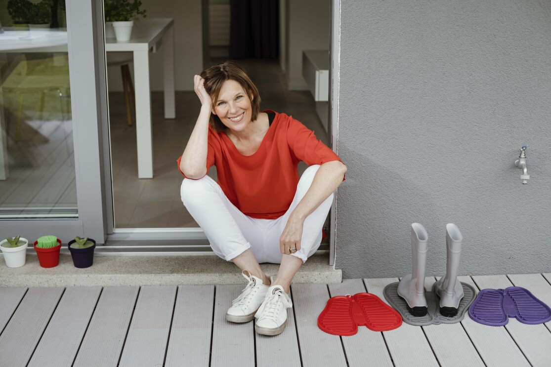 Eine Frau mit rotem T-shirt und weißer Hose sitzt in einem Türrahmen und lächelt in die Kamera.