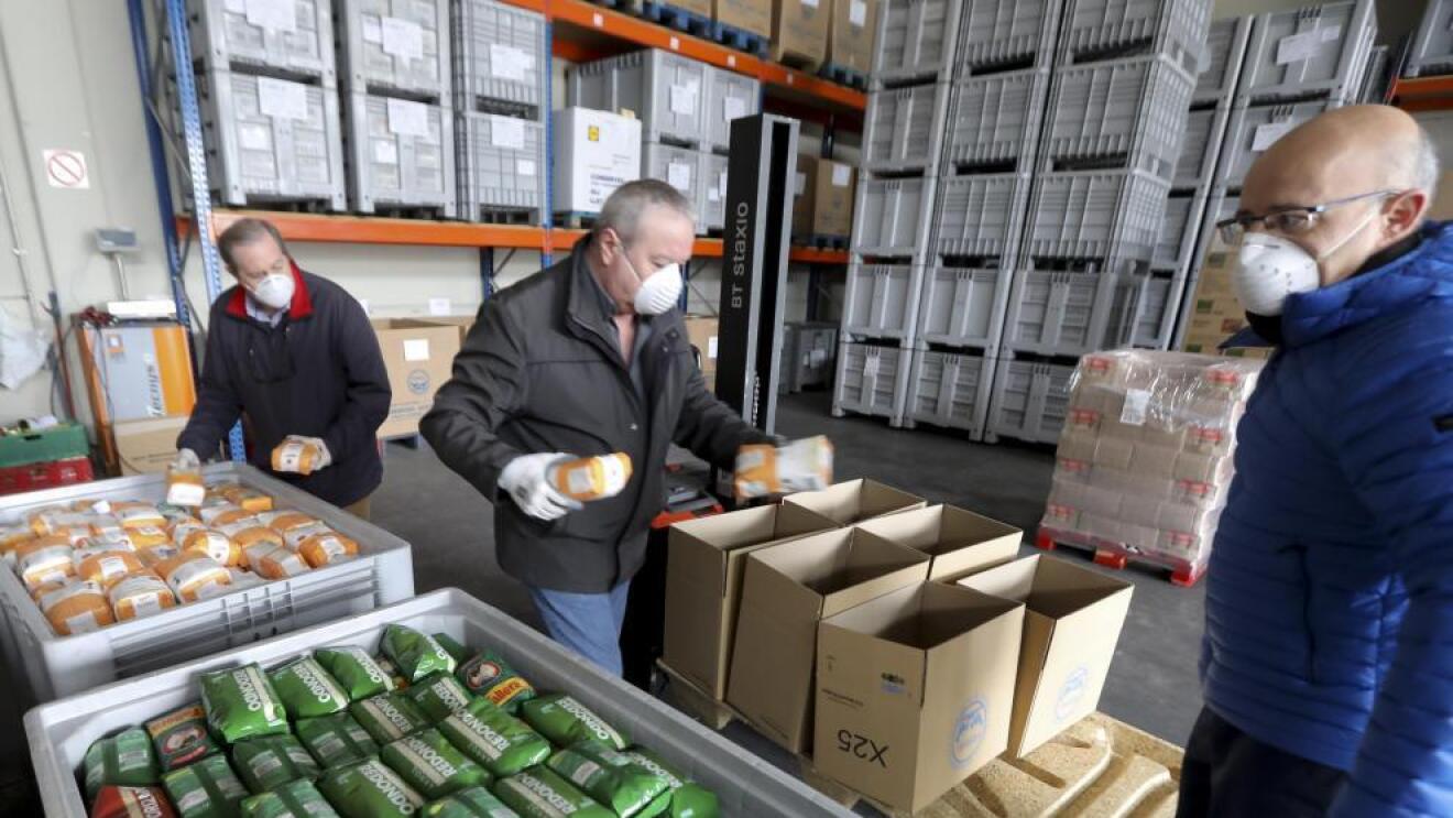 Banco de alimentos. En la parte inferior de la foto, dos cubetas grandes con paquetes de arroz y azúcar. Tres personas de mediana edad, con mascarilla, están con paquetes en las manos colocándolos en cajas de cartón. De fondo, estanterías llenas de palés de color gris.