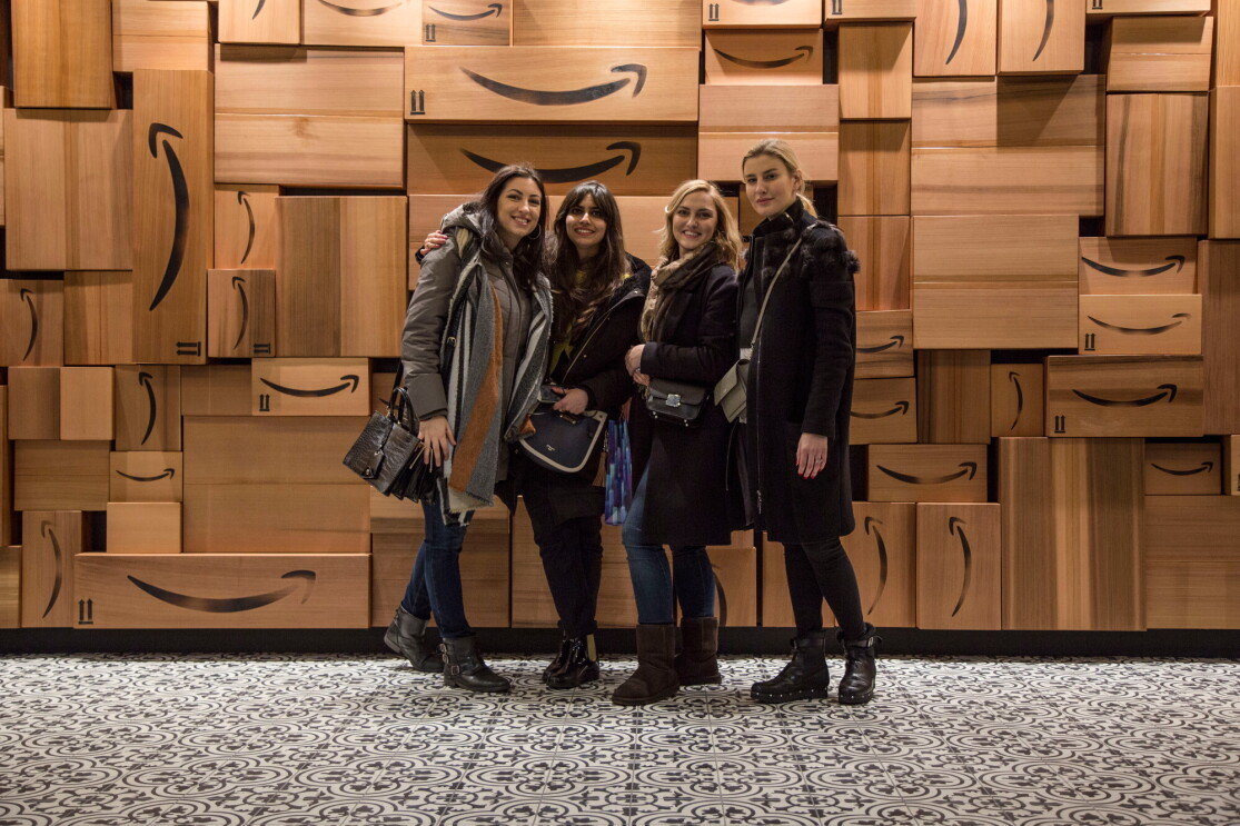 Quattro ragazze sorridono di fronte a un muro di scatole Amazon. Sono Monika, Urte, Francesca e Ankita, vincitrici dell'Amazon Design Award, quattro studentesse del Master in Interior Design dello IED Milano hanno vinto gli Amazon Design Awards, una competizione in cui si chiedeva ai corsisti di ripensare i depositi di smistamento Amazon con uno sguardo alla praticità e al comfort dei dipendenti.
