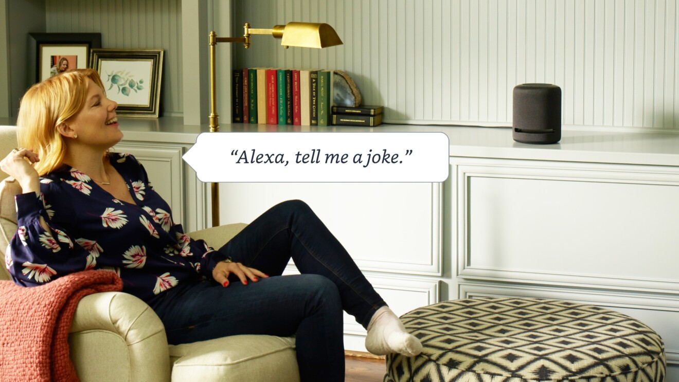 A woman sits on a chair next to Alexa in a living room. A word bubble reads, "Alexa, tell me a joke."