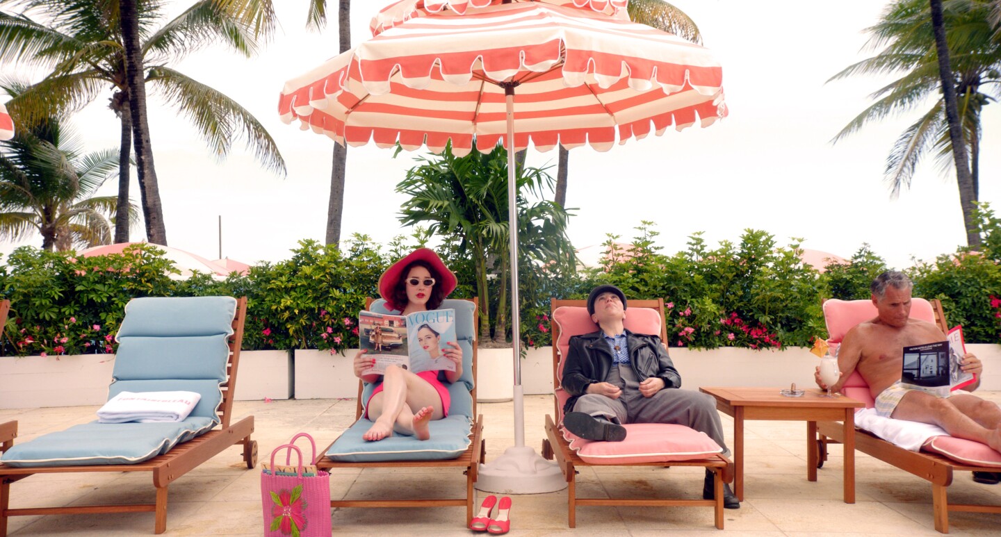 Mrs. Maisel reclines on a lounge chair, poolside, under an umbrella and reading a copy of Vogue. She's joined by Susie Myerson and a man drinking a tropical drink.