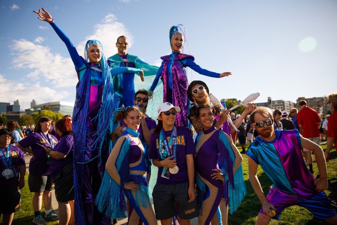 A Special Olympics USA Games athlete posts with event performers at the closing ceremony. The female athlete is wearing medals from the Special Olympics, while the performers include three stilt walkers (the two females are wearing elaborate headpieces), a juggler, and others in apparel (with lots of glitter, fringe and cutouts) that suggests a circus-themed show.