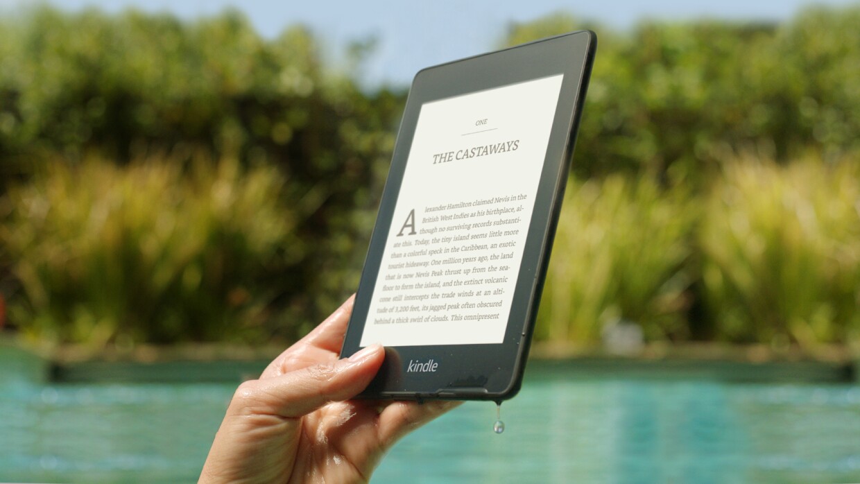 Someone holding their Amazon Kindle Paperwhite by a swimming pool. There is water dripping from the corner of the device. 