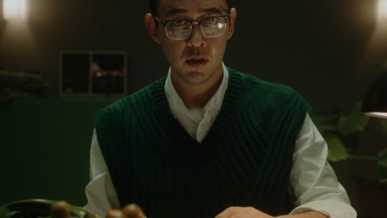Un uomo di origine asiatica siede ad una scrivania. Guarda in camera, indossa degli occhiali e ha un gilet scuro su una camicia bianca. Il resto della stanza non è ben visibile.