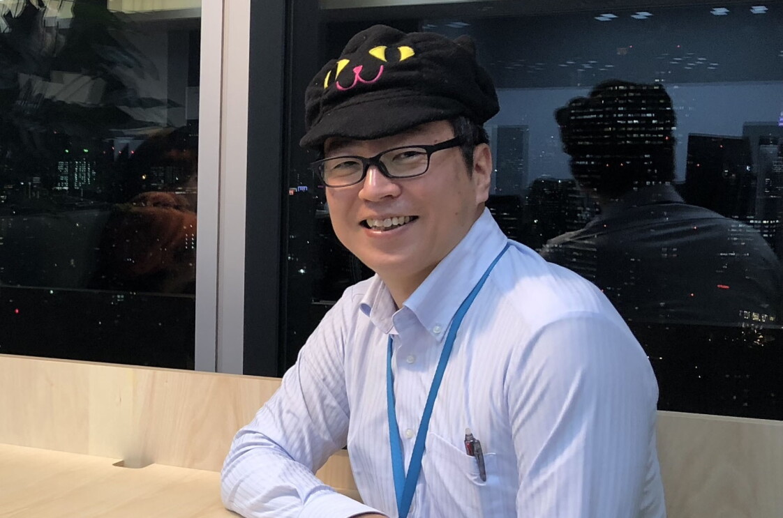黒い猫の帽子をかぶった男性が窓際のテーブルにすわりほほ笑んでいる。窓の外には夜景が見える