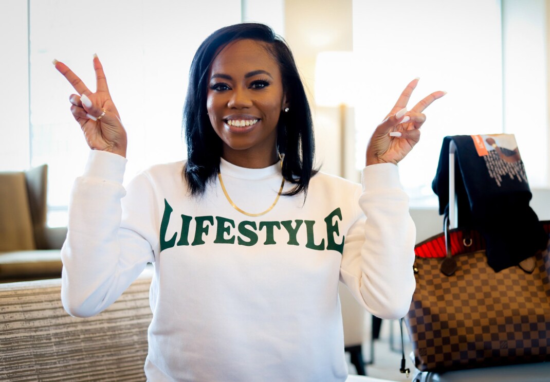 An mage of a woman smiling for a photo while making two peace signs with her hands. She is wearing a white sweatshirt that says "LIFESTYLE" on it.