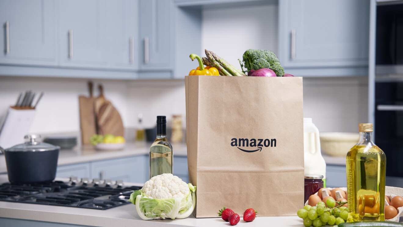 A kitchen counter top with fresh produces like strawberries, cucumber, grapes, eggs and a paper bag with the amazon label on it