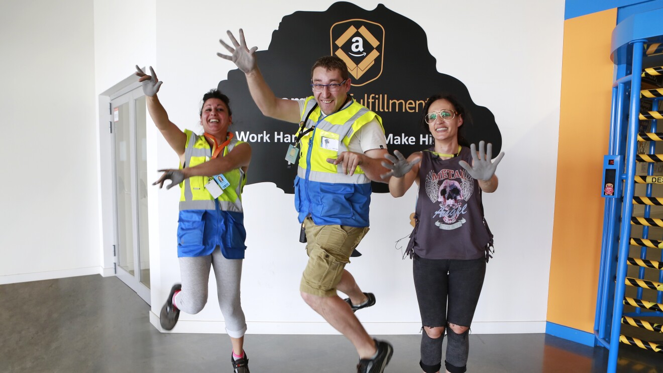 Patricia Alonso, Maribel Salcedo y Marius Dumitrescu aparecen en la entrada del centro logístico saltando y mostrando las palmas de sus manos pintadas de color gris. Están sonriendo mirando a cámara. Marius se enceuntra en medio.