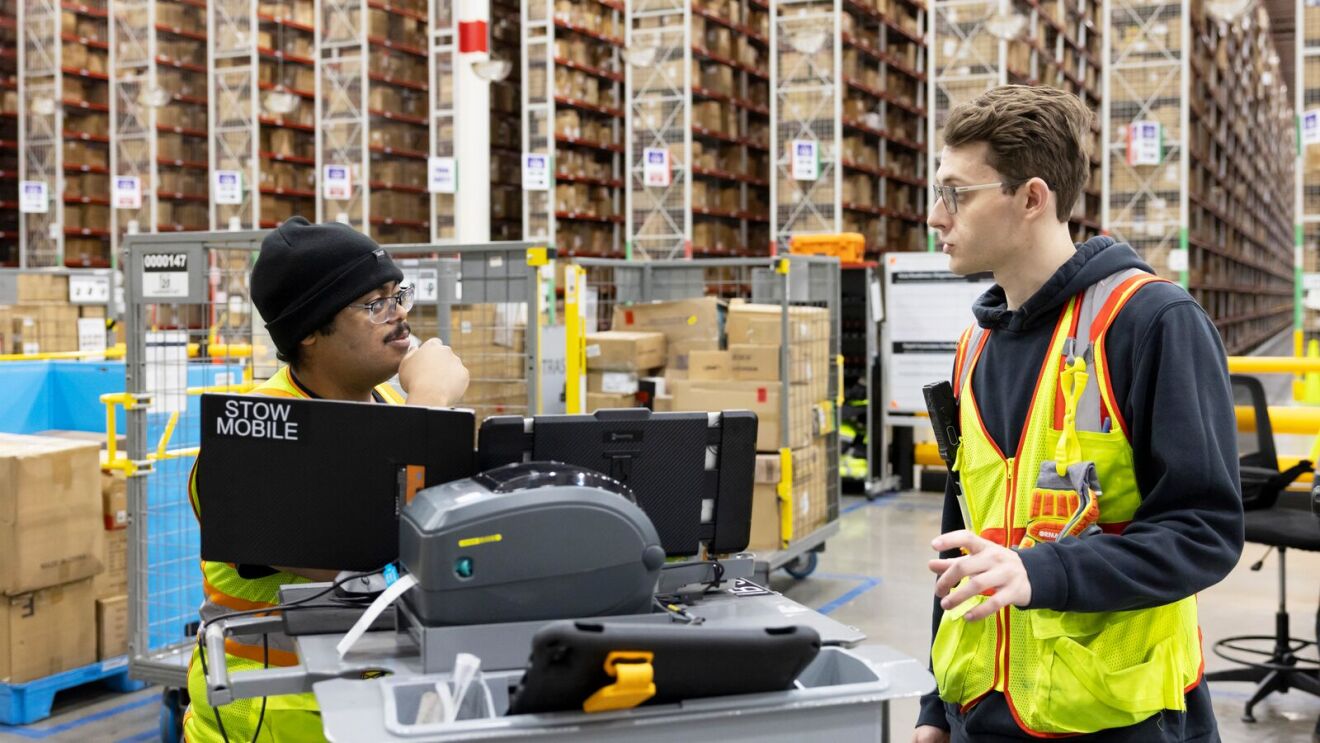 Warehouse workers in safety vests discussing at workstation with computer.