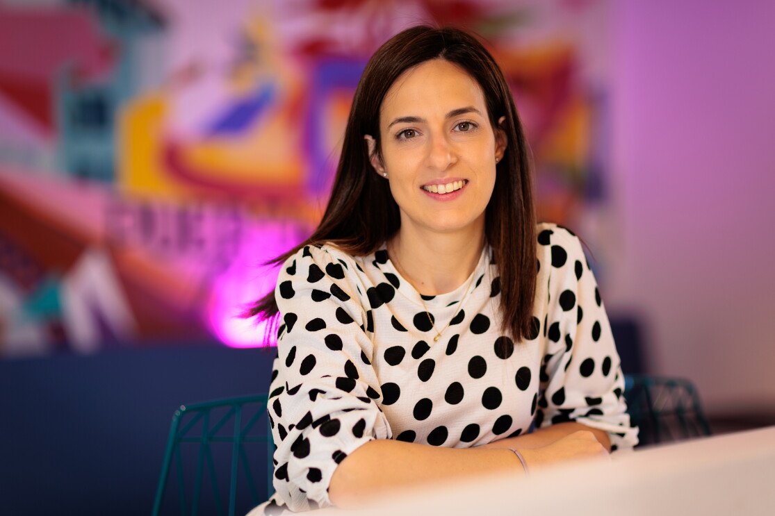 Alejandra Ossa tiene el pelo negro largo hasta el hombro. Lleva un vestido blanco de lunares negros de manga. Está apoyada  en la mesa con los brazos cruzados. Fondo de muchos colores vivos difuminados. lleva pendientes, collar y pulsera. 
