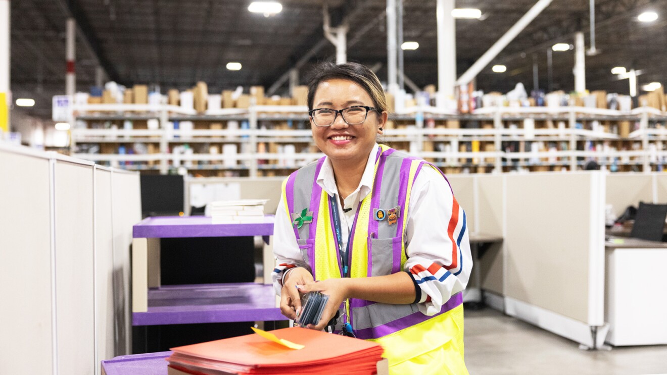 A woman wears a yellow and purple safety vest and smiles as she works in a fulfillment center.