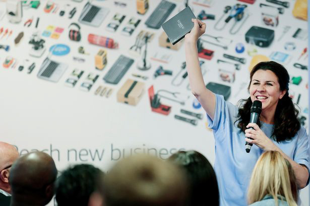 Woman with brown hair holding a mic and talking to an audience 