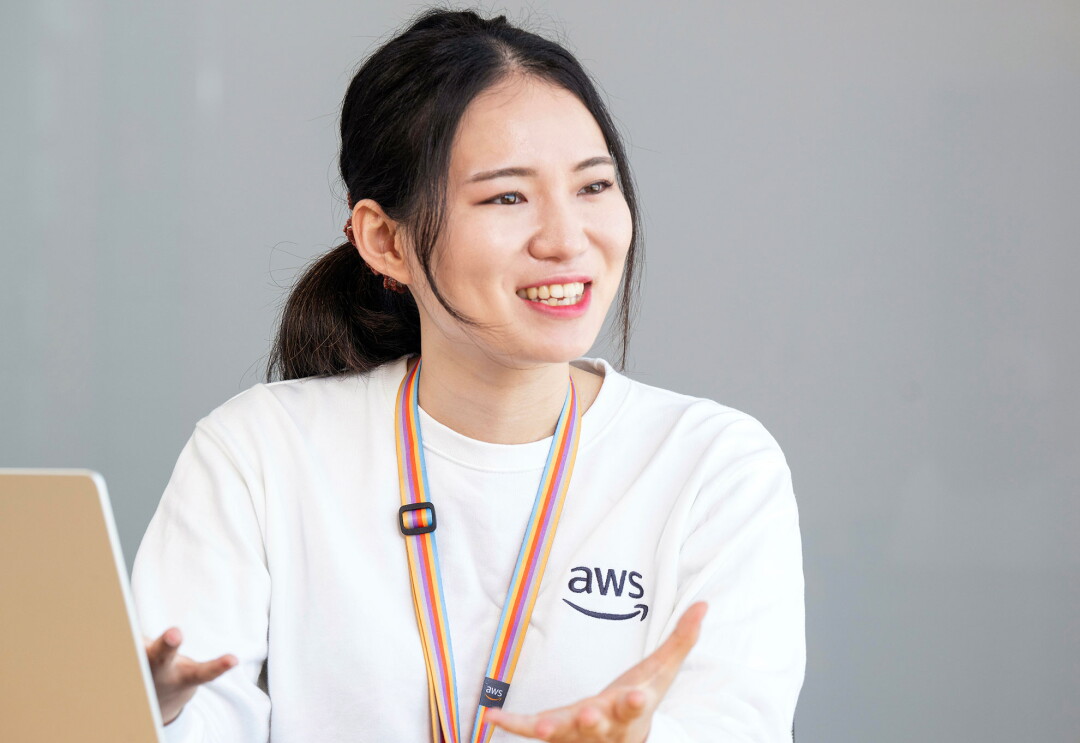 グレーの壁を背景に、AWSロゴ入りの白いスウェットを着用した女性のバストアップ。髪の毛を後ろでひとつにまとめ、カラフルな縞のストラップを首にかけている。両手のジェスチャーを交えながら話す様子。左隅に端末の背面が見える。
