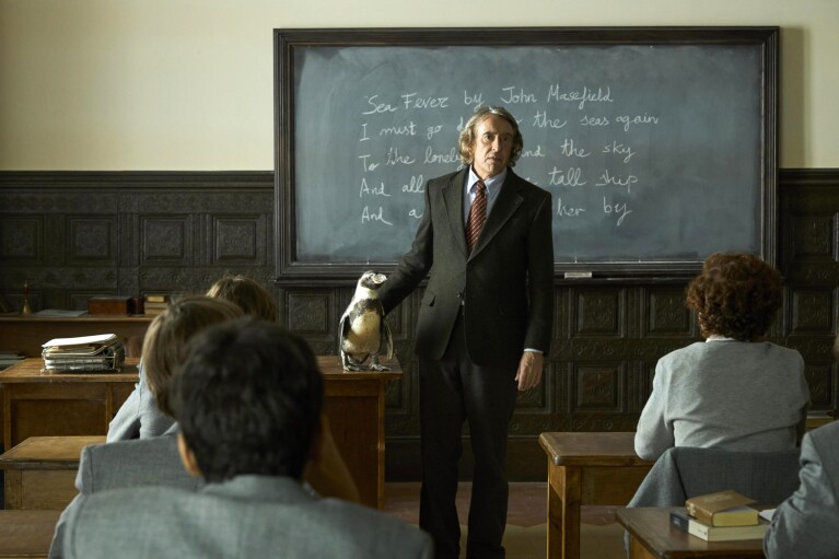 A still from The Penguin Lessons, showing a teacher with a penguin lecturing in a classroom with a poem on the chalkboard behind him