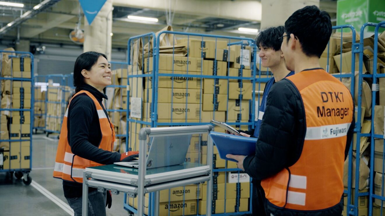 作業用ベストを着た女性と2人の男性が倉庫の中で話している。彼らの周りには多くのAmazonの段ボールが積まれたカートがある