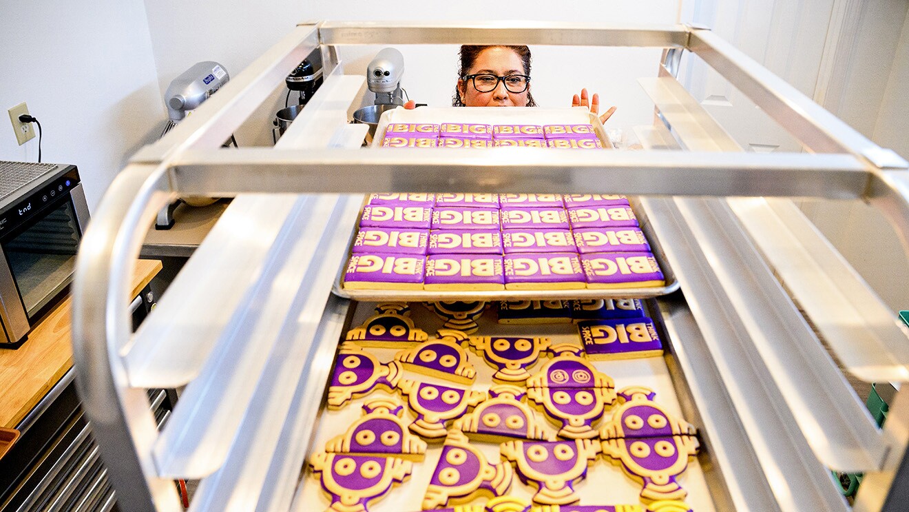 A photo of cookies decorated as AWS Think Big logos and robots cooling on cooling racks. A baker is standing behind the cookies.