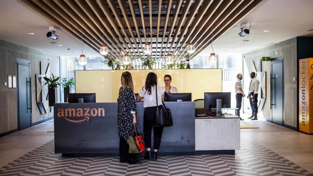 La reception degli uffici corporate di Amazon a Milano. Una donna dietroil bancone marrone, con il logo di Amazon sulla sinistra, accoglie due ospiti che rivolgono le spalle alla camera. In fondo, sulla destra, due guardie di sicurezza una di fronte all'altra parlano tra loro. 