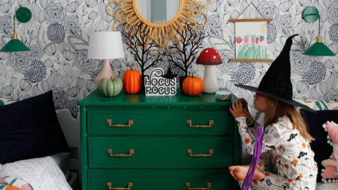 A little girl looks at an Echo Dot Kids' edition device on her nightstand while wearing a witch hat. The room is adorned in Halloween decorations.