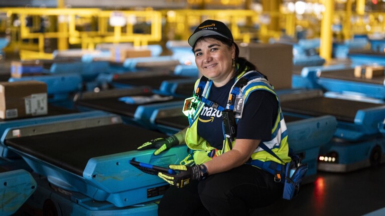 Woman in hat and safety vest posing with blue robotic equipment in Amazon facility.