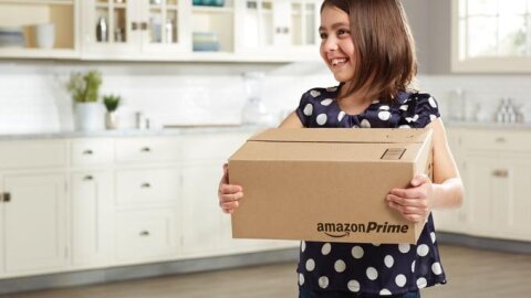 Bambina con un vestito blu a pois bianchi regge una scatola con il logo Amazon Prime, nella cucina della sua casa. 