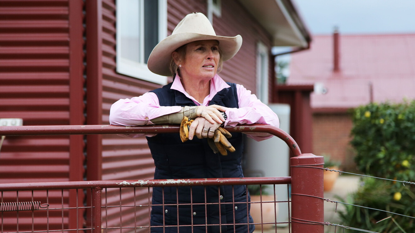 An image of a woman wearing a cowboy hat leans on a railing on on the patio in front of her red home, holding her work gloves in one hand and looking off into the distance. 