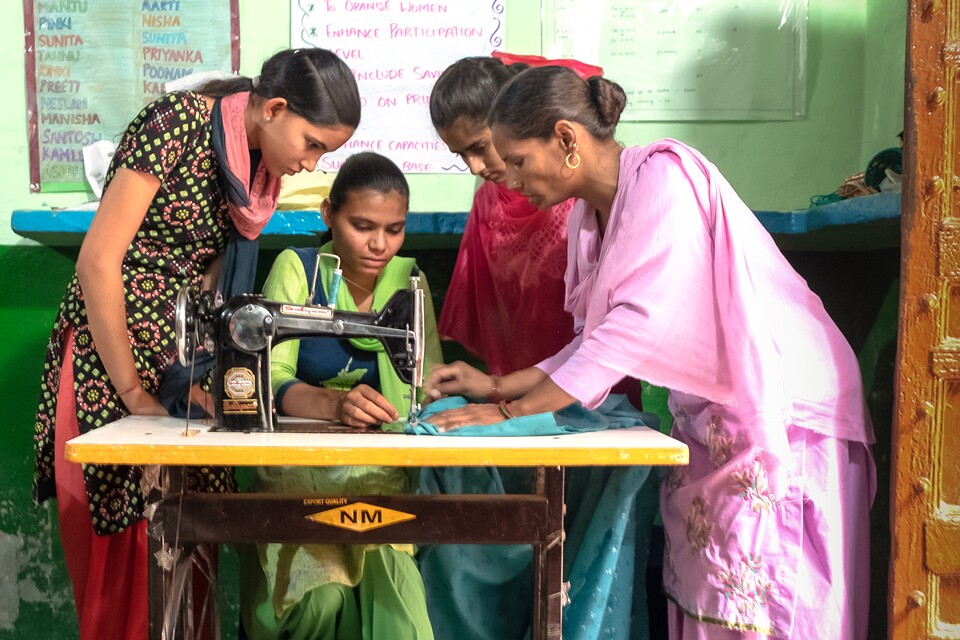 Three women stand around a woman who sits at a sewing machine.