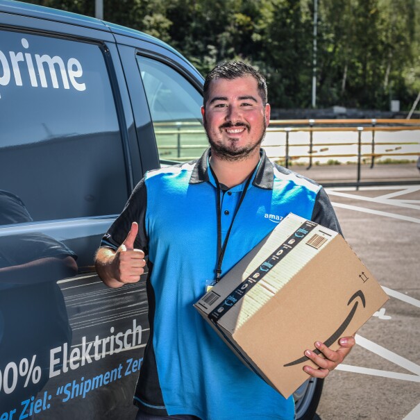 Ein lächelnder junger Mann in Amazon Shirt steht vor einem Prime Lieferfahrzeug. Er hält ein Amazon Paket in der Hand.
