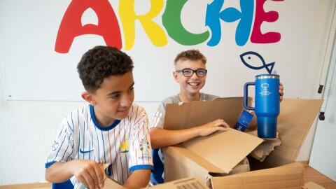 Zwei Kinder packen Pakete aus und stehen vor der Aufschrift "Arche". 