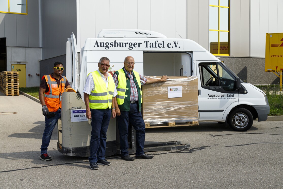 _Ein Logistikmitarbeiter mit gelber Sonnenbrille fährt via Scherenhubwagen die Paletten zum Lieferwagen der Augsburger Tafel. Mit im Bild: die beiden Vertreter der Augsburger Tafel.