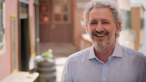 Se ve el centro de un pueblo de Italia, con las fachas de color rosa, un cartel negro con letras blancas que se ve borroso. En primer plano de la foto vemos a Sergio Panizza, de la fábrica Pesto Rossi, de salsa pesto. Sergio tiene el pelo y la barba y el bigote blanco. Está mirando a cámara sonriendo y lleva una camisa de lino azul. 