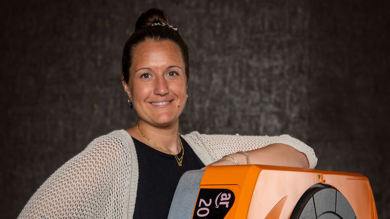 Carmen Veuthey, responsable de Operaciones en el centro logístico de Illescas. El fondo es negro, encima de un taburete hay un robot de Amazon de color naranja. Carmen está apoyada con el brazo izquierdo al robot. Va vestida con un vestido negro largo y una chaqueta crochet de punto de media manga y hasta la cintura de color beige. La mano izquierda la tiene en la cintura. Va con collares de oro, pendientes y un moño alto. 