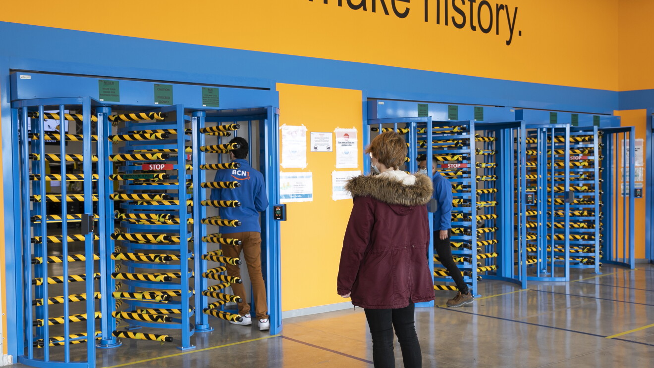 Se han instaurado prácticas específicas para garantizar la distancia de seguridad entre personas en nuestros edificios. En una entrada de un centro logístico hay dos personas entrando por el rodillo de fichar. Hay tres personas entrnado manteniendo la distancia de seguridad. Todos están de espaldas. Los dos chicos ya entrando y una chica con un abrigo y mascarilla de espaldas esperando en una línea amarilla que hay en el suelo.
