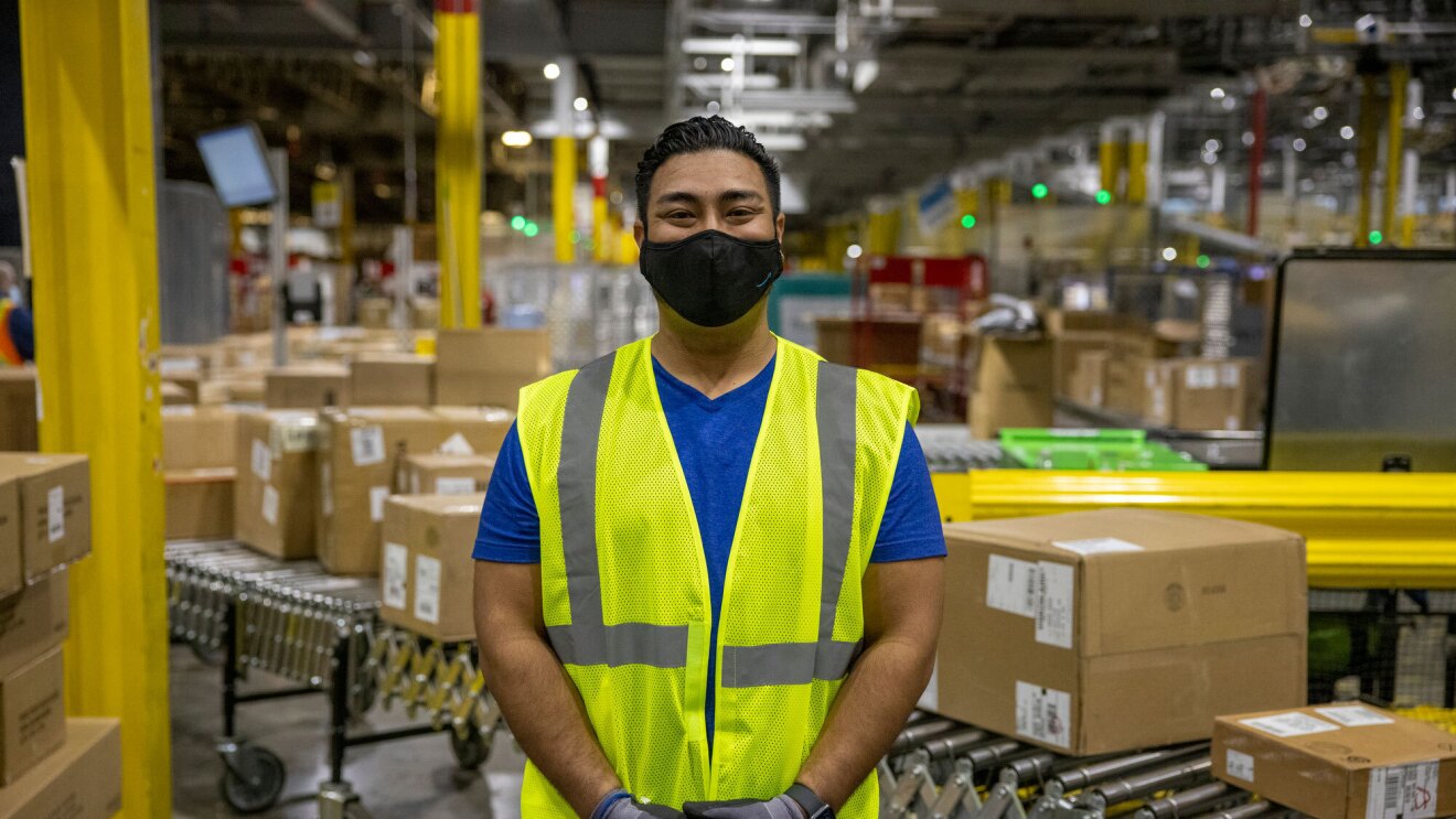 A male employee wearing a face mask and a yellow safety vest stands on the floor of an Amazon fulfillment center, looking at the camera. Boxes are on a conveyor belt behind him.