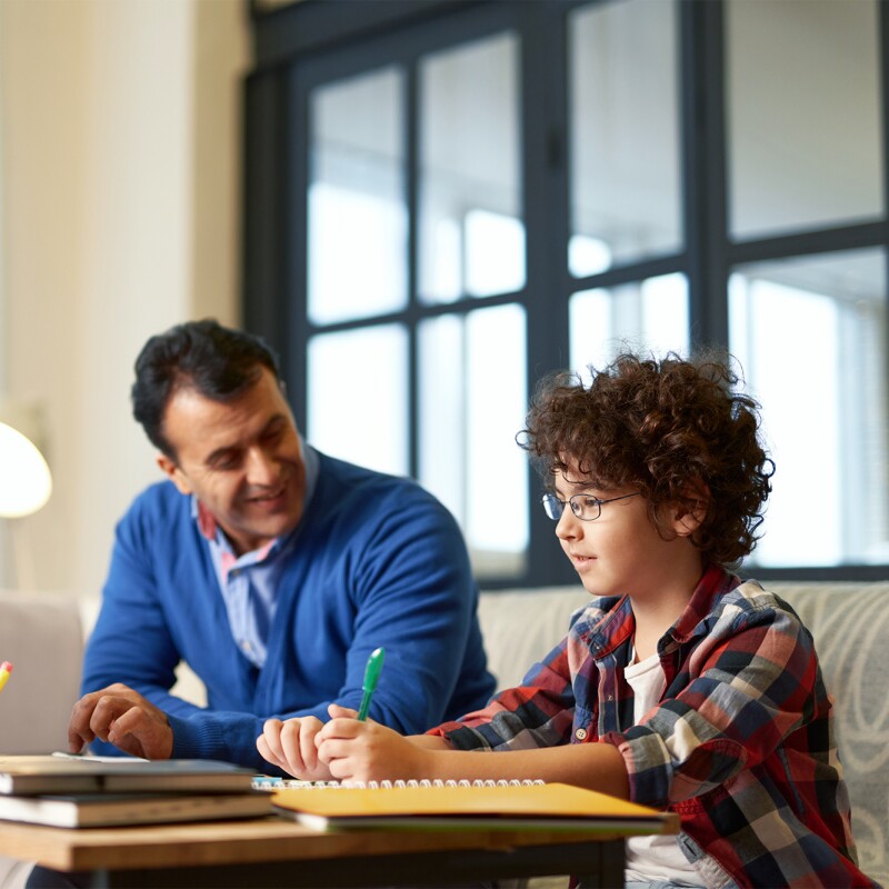 Um homem sentado em frente a um computador e ao lado de um menino, que escreve em um caderno. Eles estão sentados em um sofá