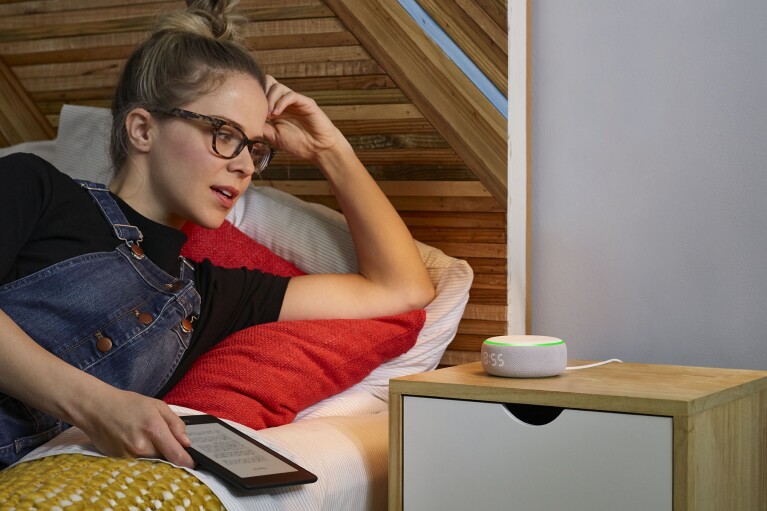 Woman wearing glasses lays on a bed as she looks at and speaks to an Amazon Echo device on a nightstand.