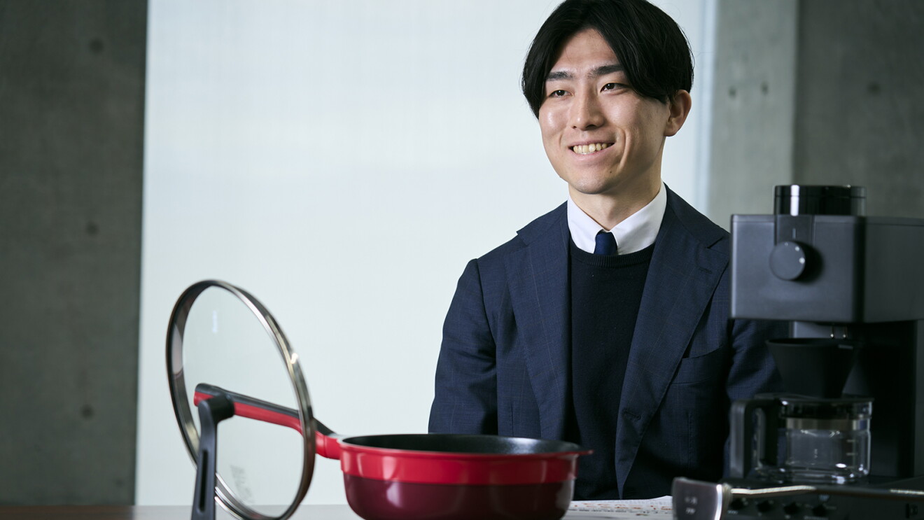 コンクリートの無機質な壁の室内で、腰掛けて話すスーツ姿の男性。台の上に赤いフライパンとガラスのふた、黒いコーヒーメーカーが置かれている。