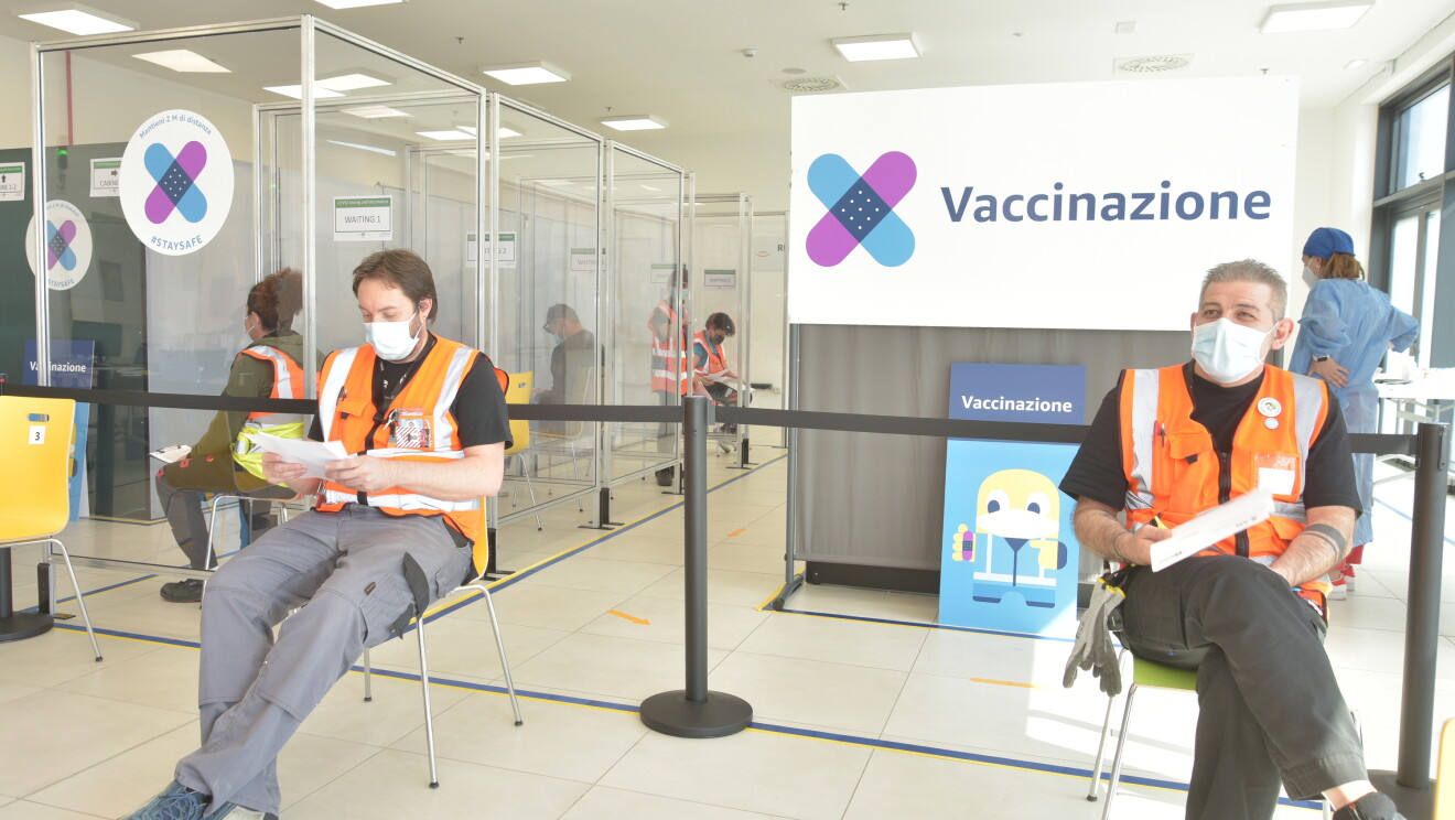 Due dipendenti Amazon attendono il loro turno per ricevere il vaccino contro il covid-19 in un'area dedicata di un sito Amazon. Sulla destra, alle spalle di uno dei due, si vede un cartellone bianco con la scritta 'Vaccinazione' e due cerotti colorati che si incrociano