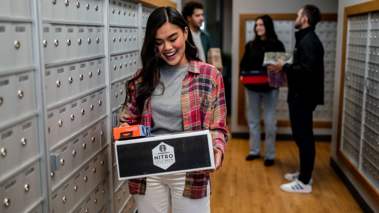 An image of a woman standing in a mailroom smiling while looking down at a package she is holding. The package has branding on it for Nitro cold brew coffee.