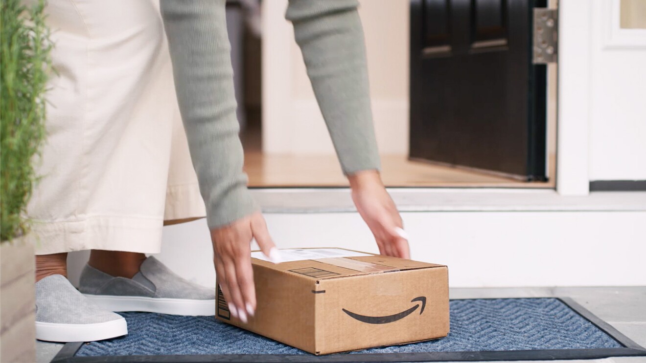 A woman in gray slip-on sneakers and white wide-leg pants bends down to pick up the Amazon package on her doorstep. 