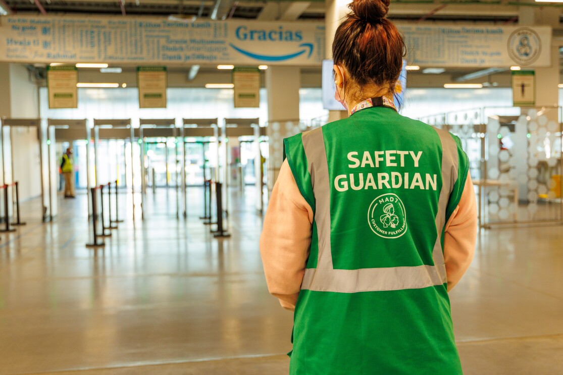 En la entrada de un centro logístico de Amazon aparece un Safety Guardian de espaldas. Es una mujer con una coleta recogida alta. La persona lleva mascarilla y un colar del que lleva el badge colgado. Tiene un chaleco de color de color verde con las letras blancas Safety Guardian MAD7.