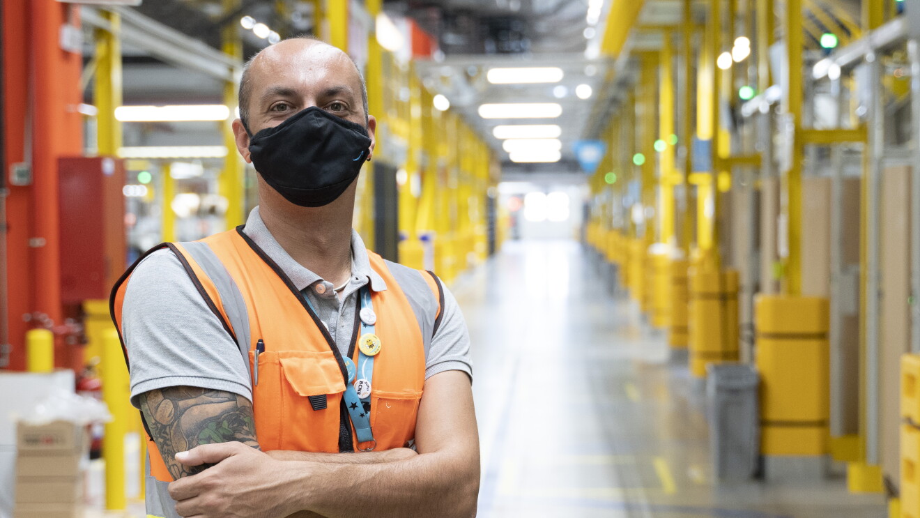 Una foto a mezzo busto di Oreste Tagliaferri con alle spalle il corridoio di un centro logistico di Amazon. Oreste indossa una mascherina nera in tessuto, un giubbino di sicurezza. Ha le braccia conserte e sul suo braccio destro si intravede un tatuaggio.