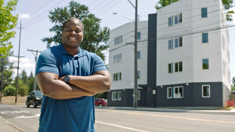 A photo of a man crossing his arms in front of an apartment building.