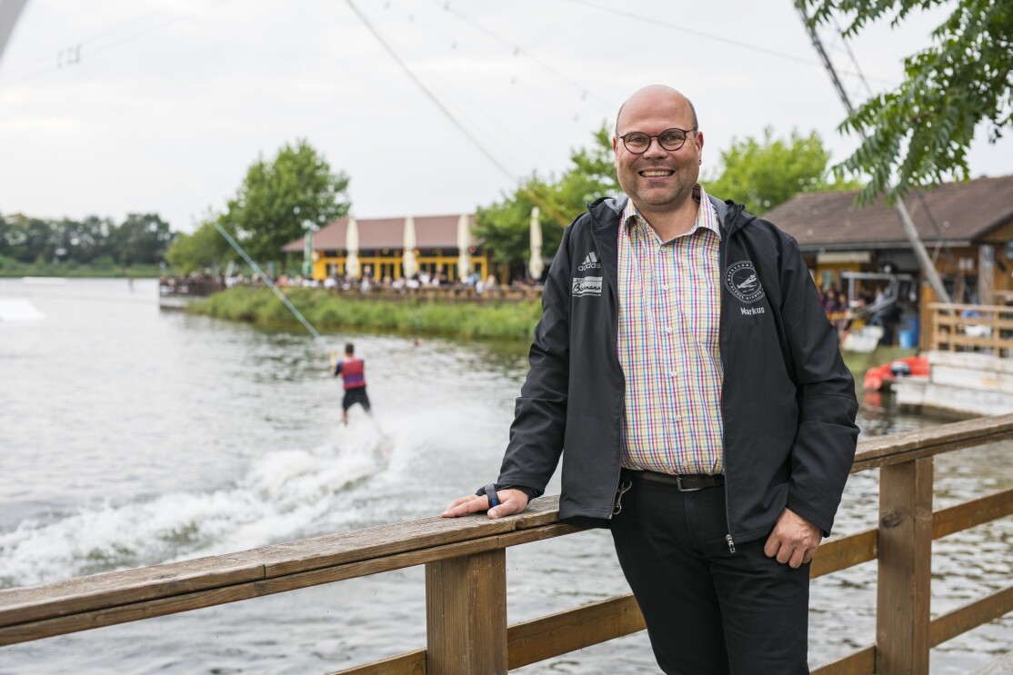 Ein man im karierten Hemd steht vor der Wasserski-Anlage und lächelt in die Kamera.