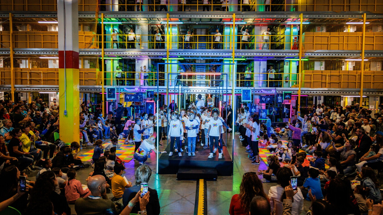 Panorámica del evento. Dentro de un centro logístico, con las estanterías de fondo y las barandillas de color amarillo, aparece una pasarela de color negro ambientada con luces de neón. Un gupo de niños con jeans y camisetas blancas del evento con pinceldas de colores están realizando una correografía con las manos en la cabeza. Los trabajadores de Amazon que son los padres de los niños que están bailando en la pasarela están mirando y sacando fotos con el teléfono. 