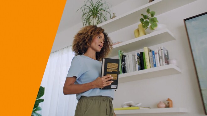 An image of a woman holding a book while talking to Alexa. There is an orange stripe on the left side. 