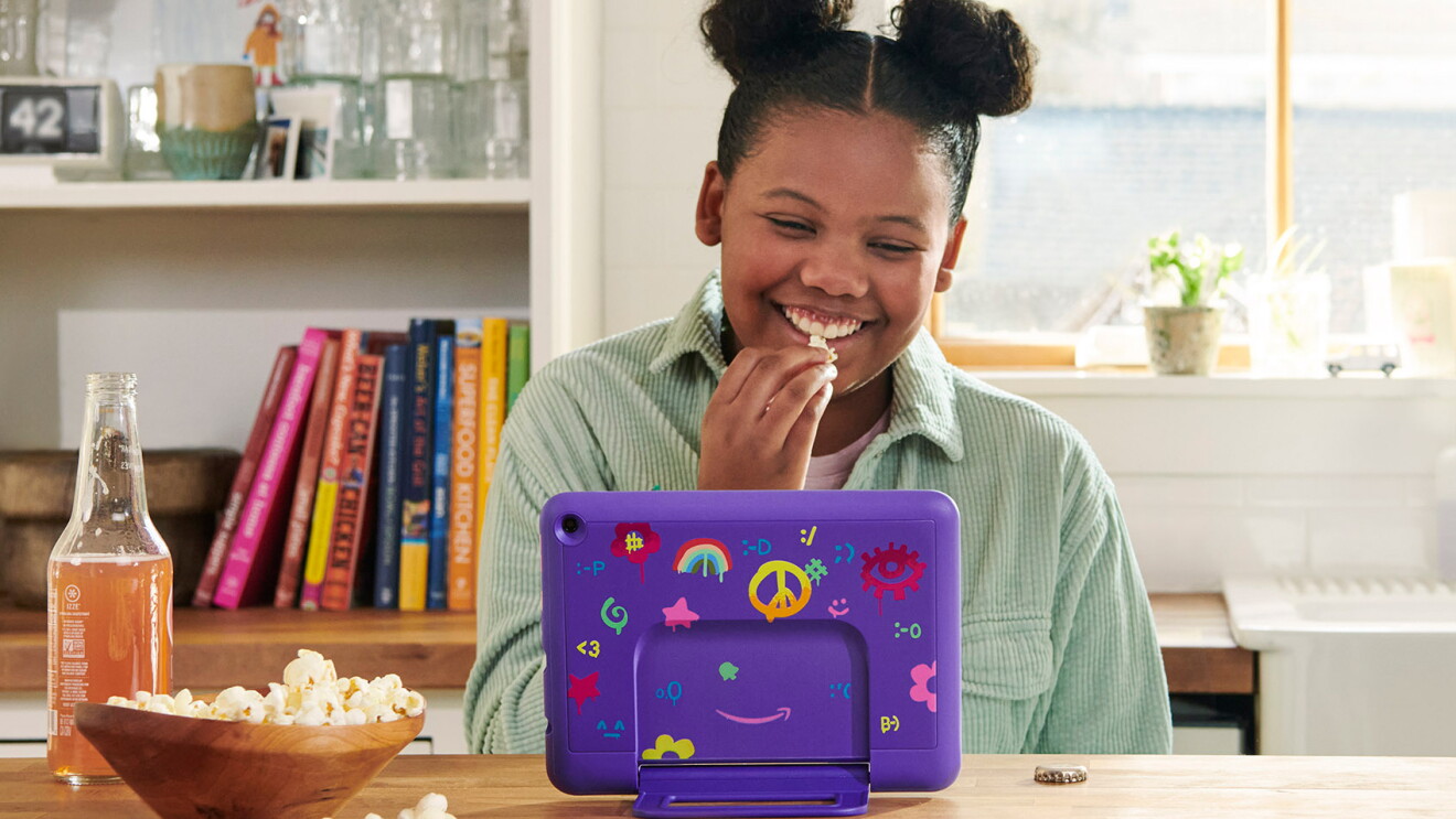 A young girl smiles as she watches her Fire Kids tablet that is protected with a purple case decorated with stickers. She is eating popcorn and has an orange soda.